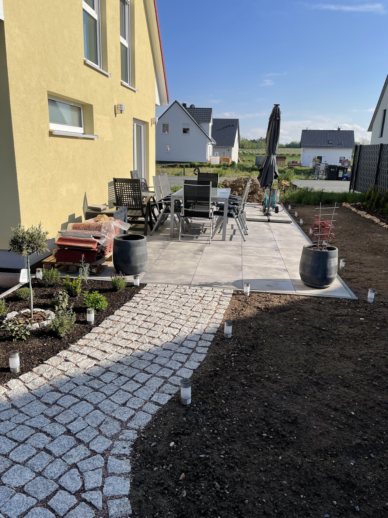 Gartenpatio am gelben Haus mit Esstisch, Stühlen, Sonnenschirm und Pflanzkübeln, gepflasterter Weg.