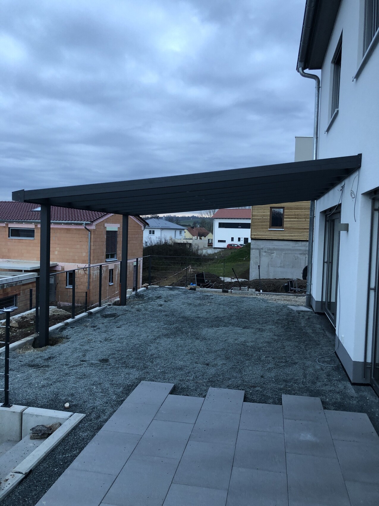 Terrasse mit schwarzer Metallpergola vor weißem Haus; Kiesboden und Terrassenplatten im Vordergrund.