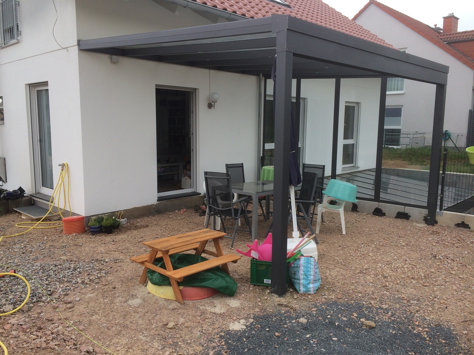 Moderne dunkelgraue Pergola über Terrasse mit Esstisch und Stühlen vor weißem Haus.