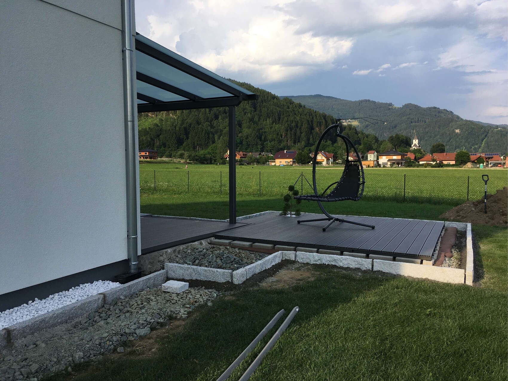 Moderne Terrasse am Haus mit schwarzem Hängesessel auf Holzdeck vor grüner Wiese und Berge.