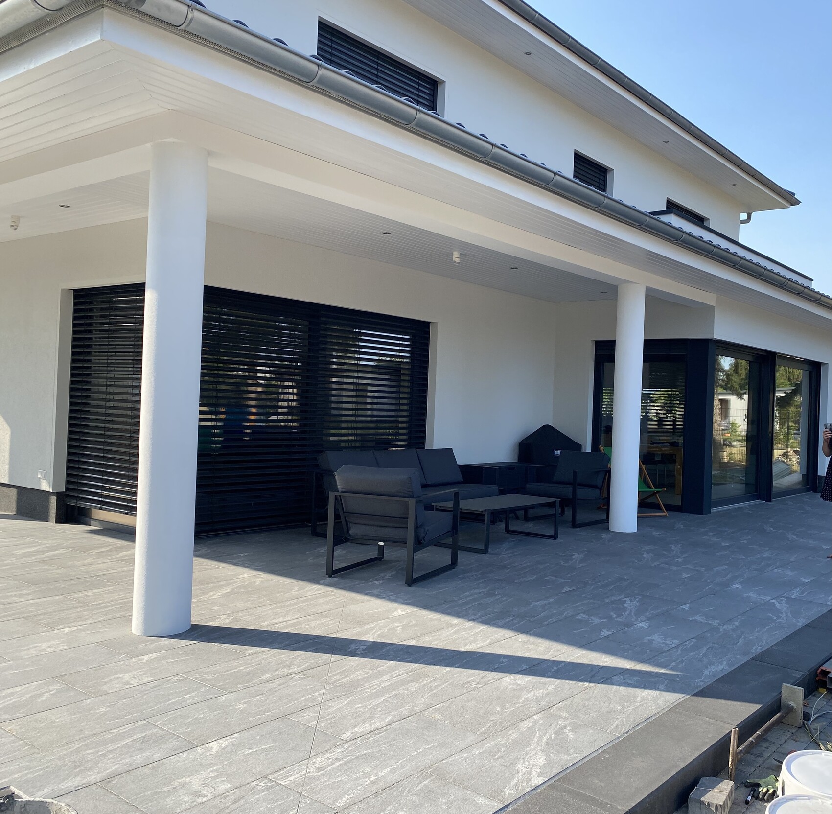 Moderne weiße Villa mit überdachter Terrasse, schwarze Lounge-Möbel und Glasschiebetüren.