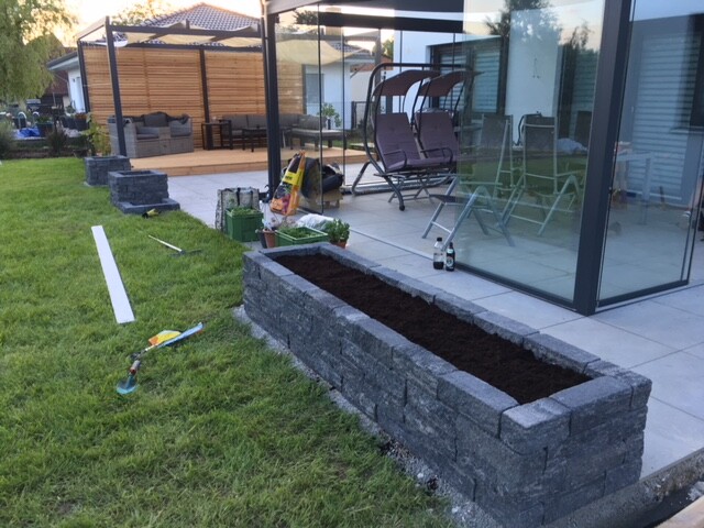 Gartenbereich mit rechteckigem Beet aus Steinblöcken neben Terrasse, Erde eingefüllt.