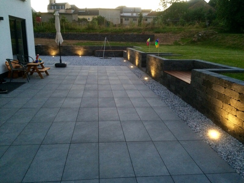 Gepflasterte Terrasse aus grauen Quadraten, linke Outdoor-Möbel, beleuchtete Steinmauer.