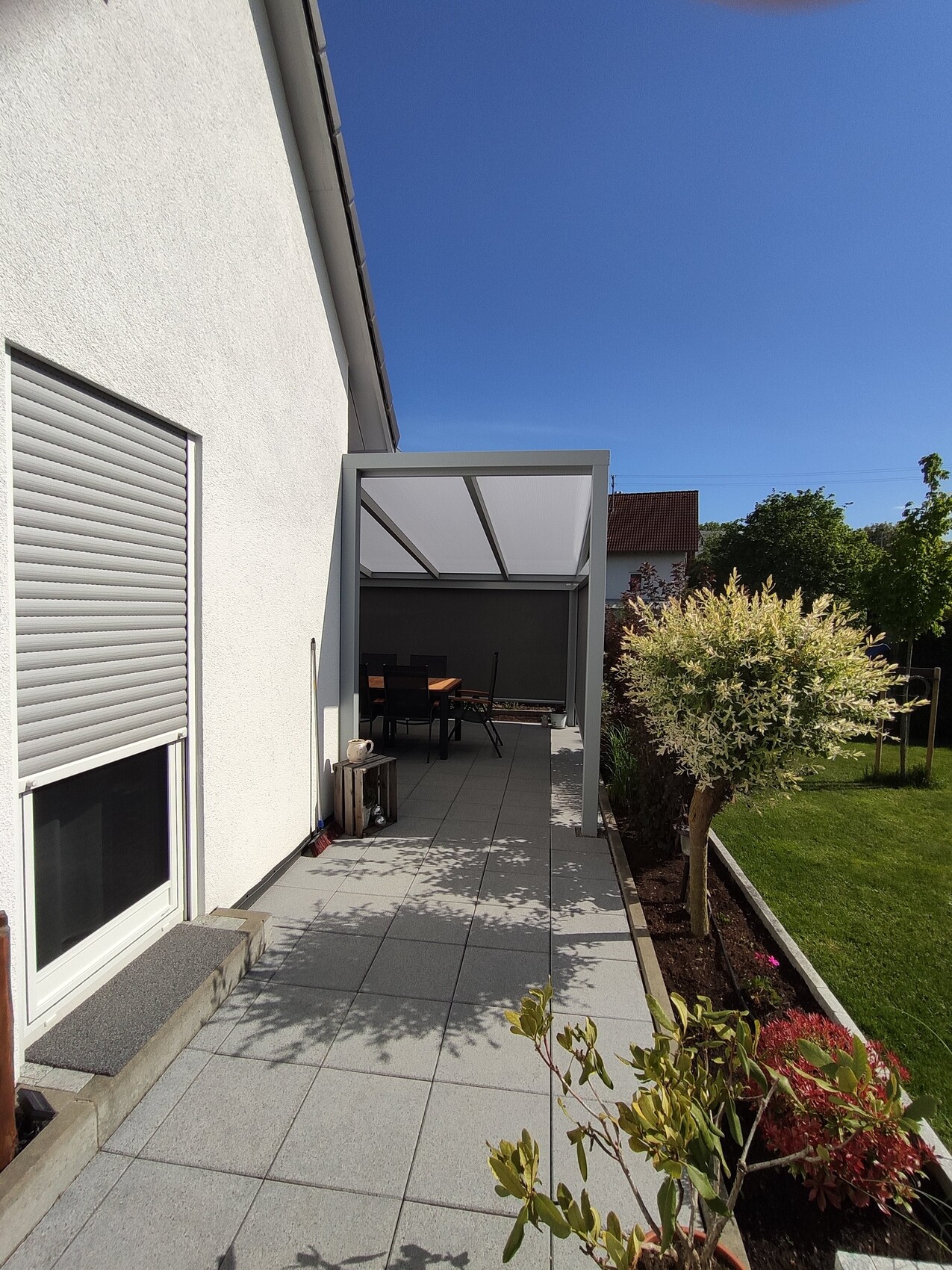 Weißes Haus, überdachte Terrasse mit Esstisch und Stühlen, gepflasterter Boden, Garten mit Baum.