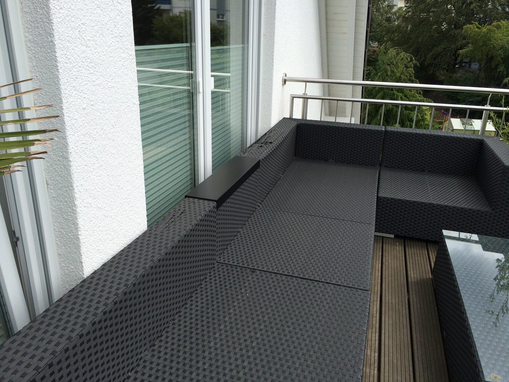 L-förmige dunkle Rattan-Sitzgruppe auf Holzboden-Balkon vor Glasfenstern und grüne Aussicht