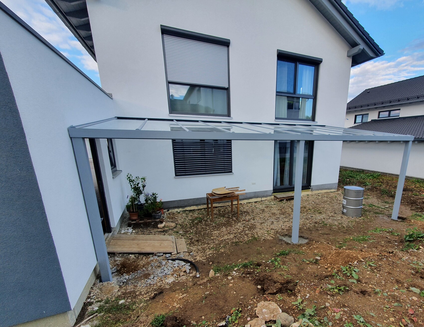 Weiße Hausfassade mit grauer Glasdach-Pergola im Hinterhof, Boden aus Erde und Kies, Pflanzkübel.