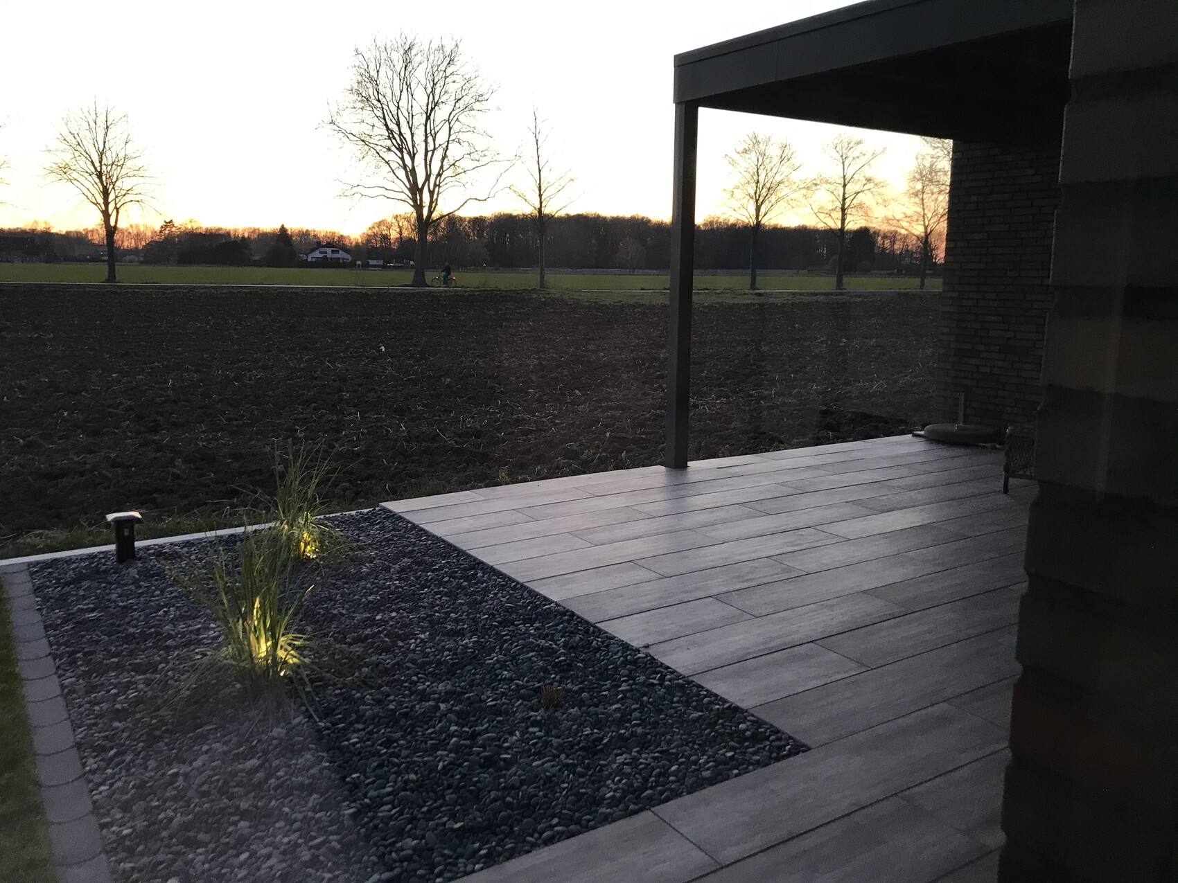 Moderne Terrasse mit grauen Fliesen, Kiesbeet, Bodenleuchten; Blick über Felder zum Sonnenuntergang.