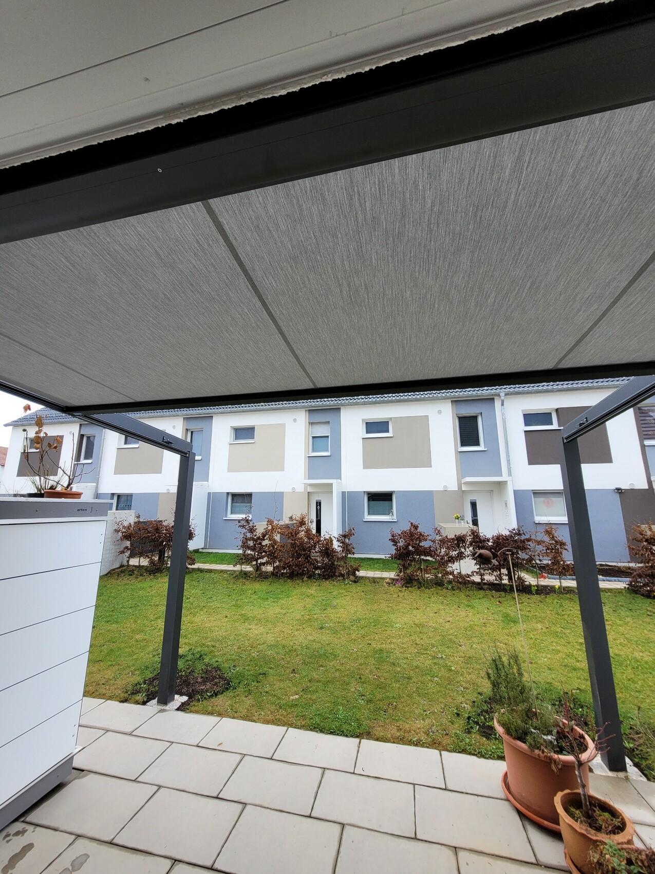 Überdachte Terrasse mit grünem Rasen, Sträuchern und modernen Reihenhäusern dahinter.