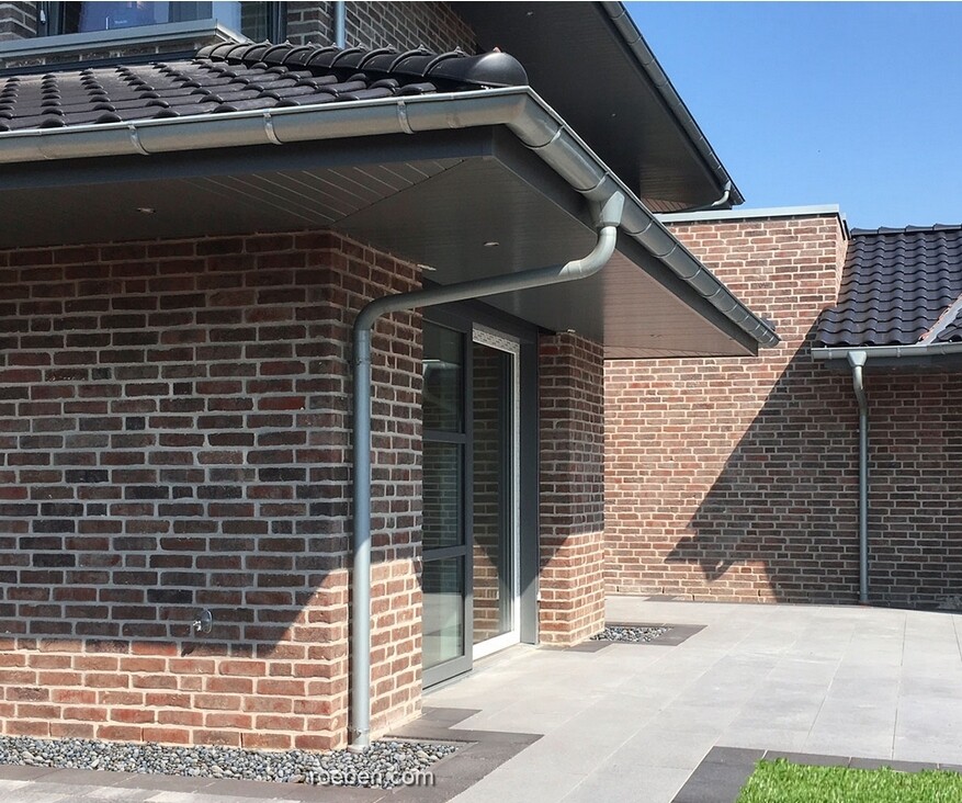 Moderne rote Ziegelwand eines Gebäudes mit Dachrinne, Fallrohr und Glastür am sonnigen Patio.