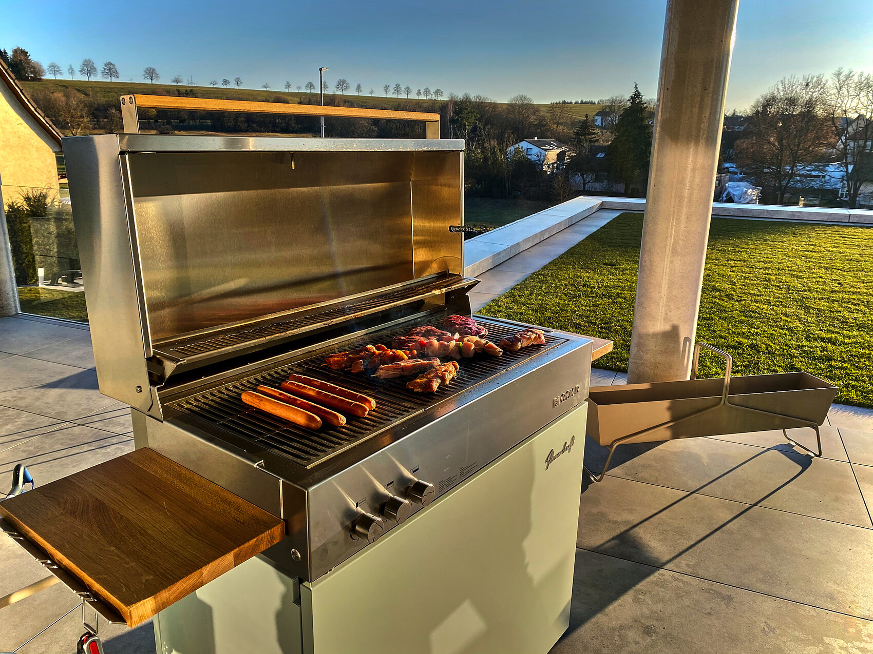 Außen-Gasgrill auf Terrasse, offen; Würstchen und Fleischspieße brutzeln, Gartenblick.