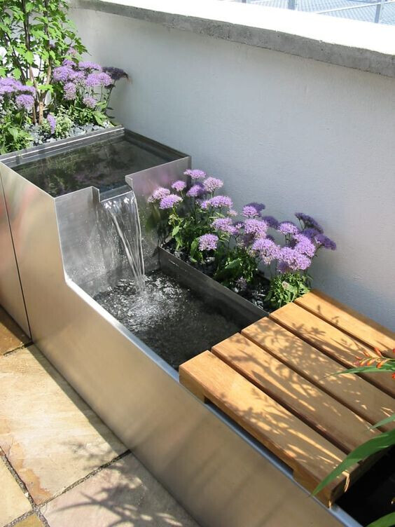 Moderne Edelstahl-Wasserschale mit Wasserfall, umgeben von violetten Blumen und Holzbänken.
