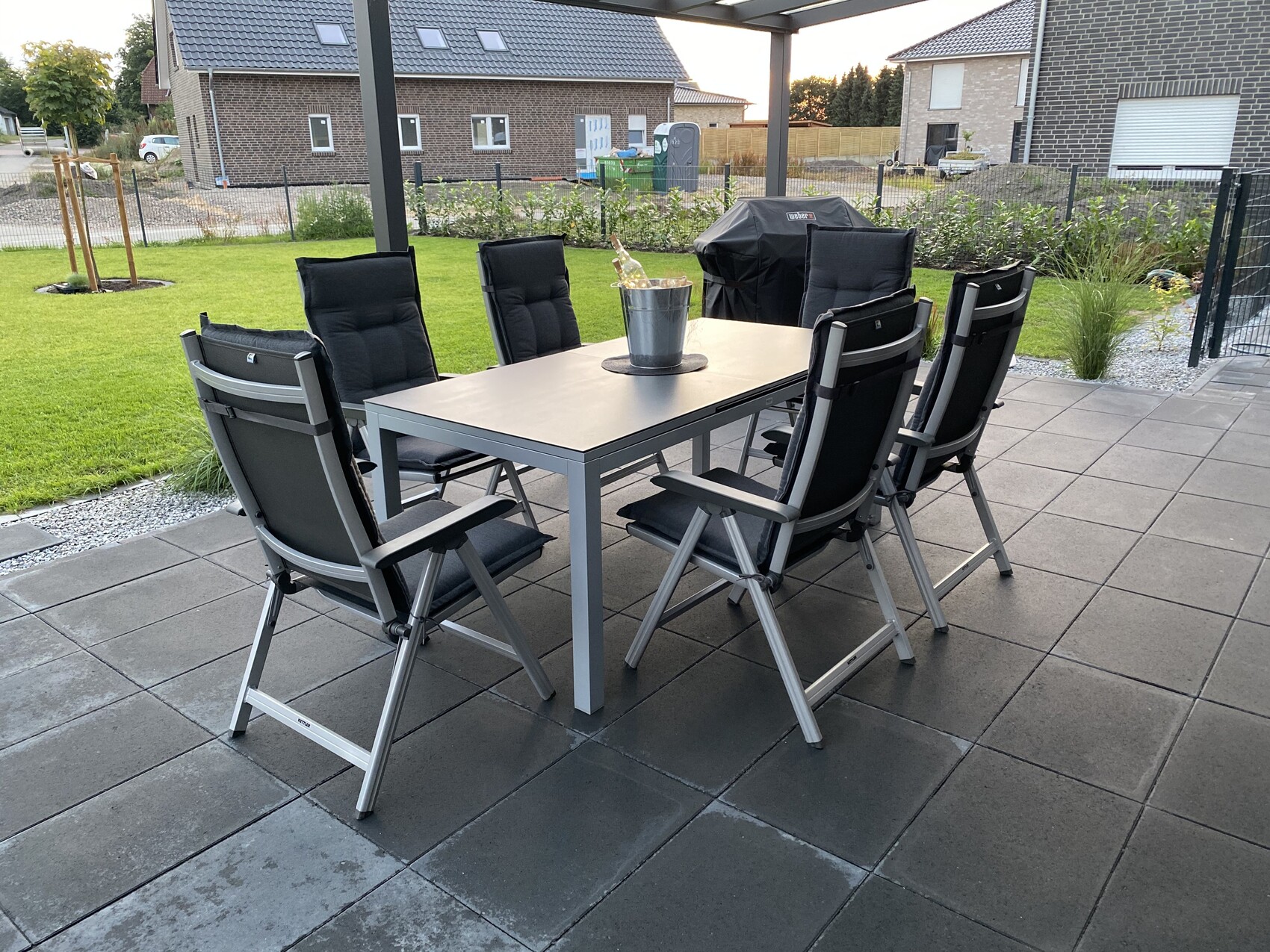 Garten-Tisch mit mehreren schwarzen Stühlen auf Terrasse, Grillabdeckung im Hintergrund.