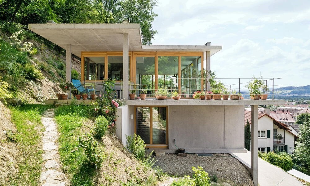 Modernes Betonhaus auf Hang mit Glasfront, Balkon voller Topfpflanzen und Aussicht.