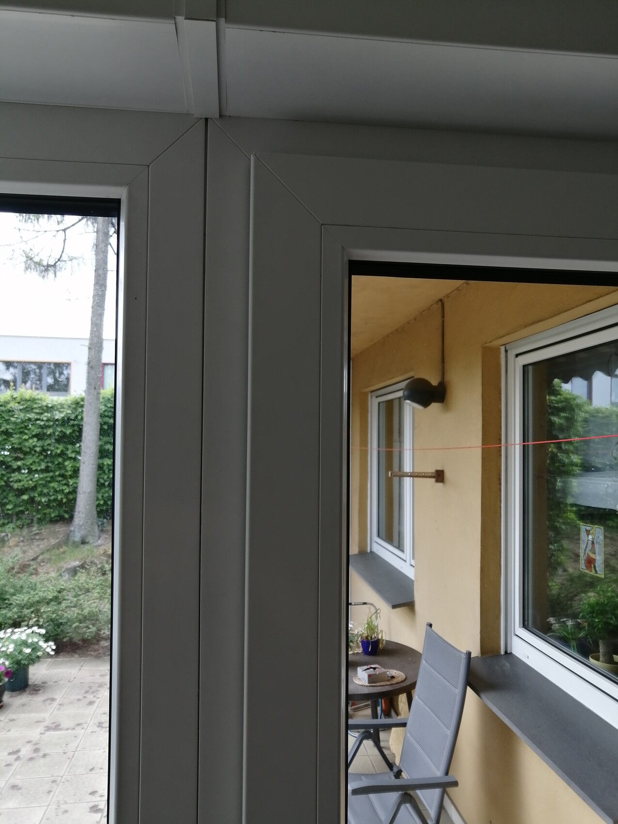 Blick durch Glasfenster auf Balkon: gelbe Wand, Tisch, Stuhl, rote Wäscheleine, Garten.
