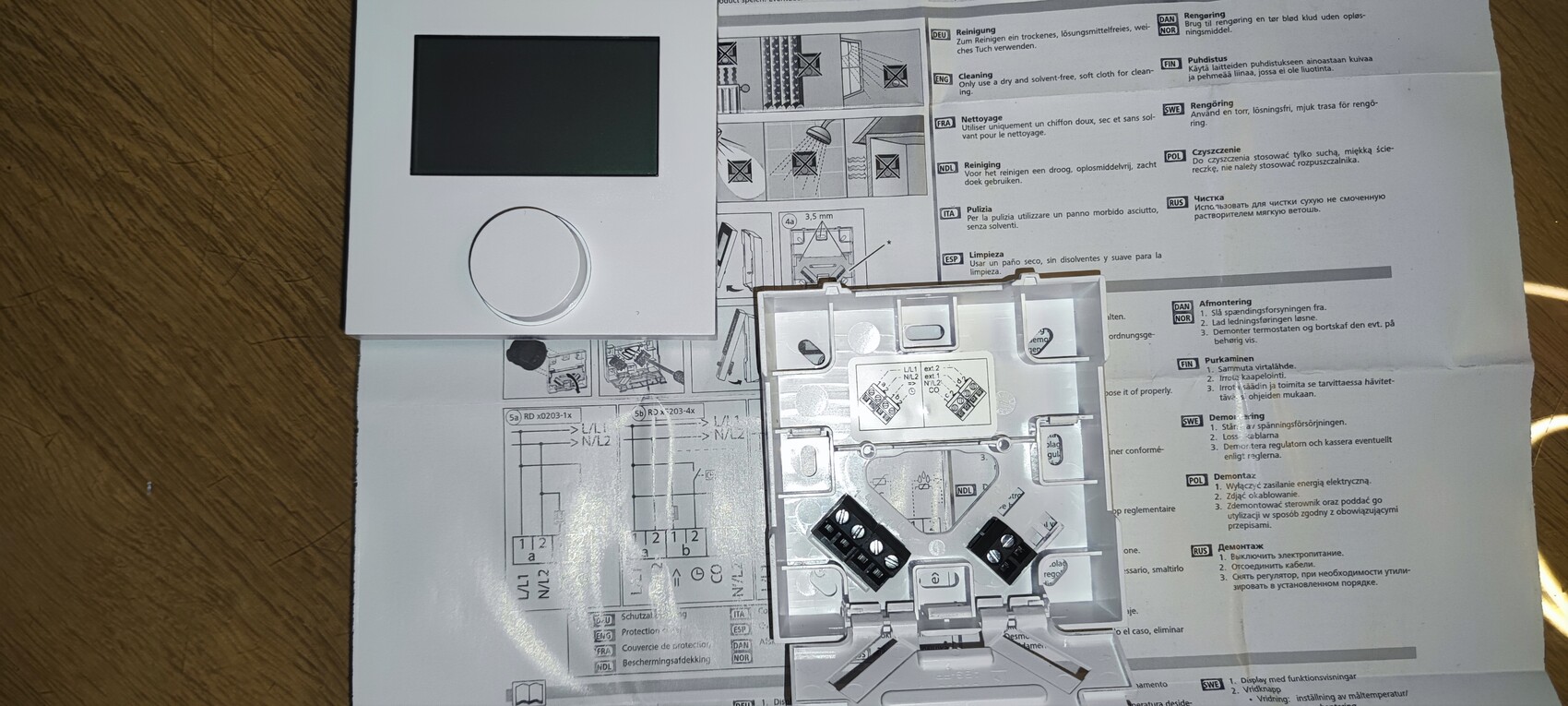 Weiße Wandbedieneinheit mit schwarzem Display und großem runden Knopf liegt auf Anleitung.