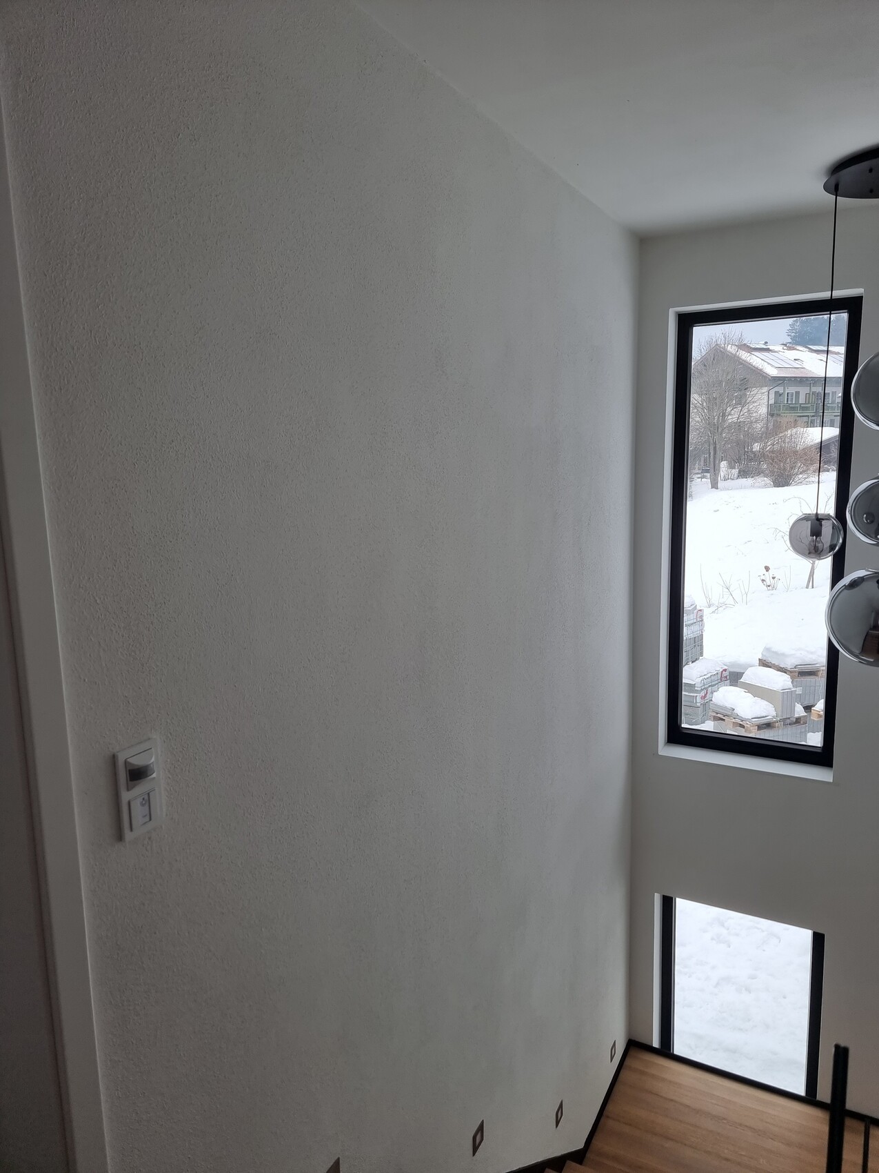 Helles Treppenhaus mit hohen Wänden, schmalem Fenster; Blick auf winterliche Außenlandschaft.
