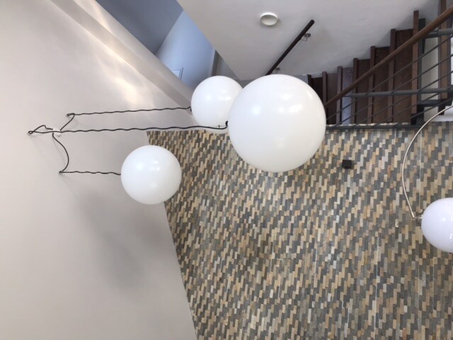 Innenraum mit Treppe; mehrere weiße Ballons hängen an Drahtseilen über einer gemusterten Wand.