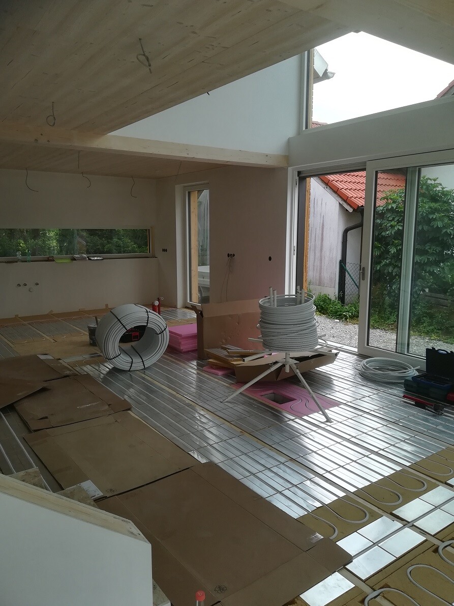 Baustellenraum mit Fußbodenheizungseinbau: Heizungsrohre, Rollen und Glasfront nach draußen.
