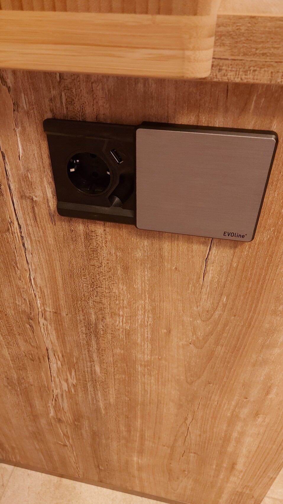 Schuko-Steckdose mit USB-Port und grauer Edelstahlabdeckung an einer Holzwand.