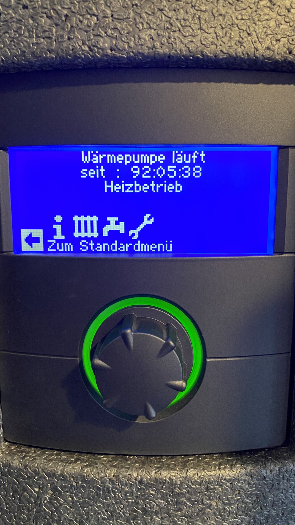 Wärmepumpe läuft seit 92:05:38, Heizbetrieb, blaues Display, grün beleuchteter Drehknopf.
