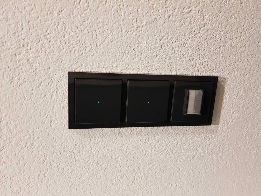 Wand-Schalterpanel: zwei schwarze Lichtschalter mit grüner LED, rechts Dimmer mit weißem Schieber.