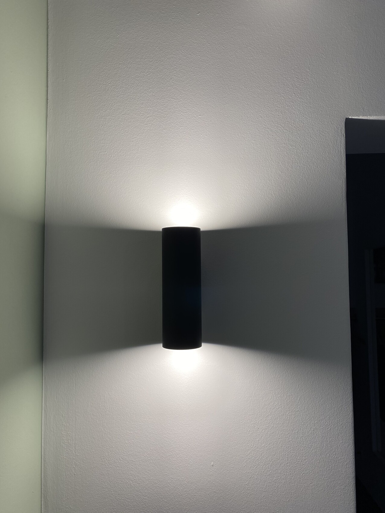 Schwarze, zylindrische Wandleuchte an einer weißen Wand; Licht strahlt oben und unten.