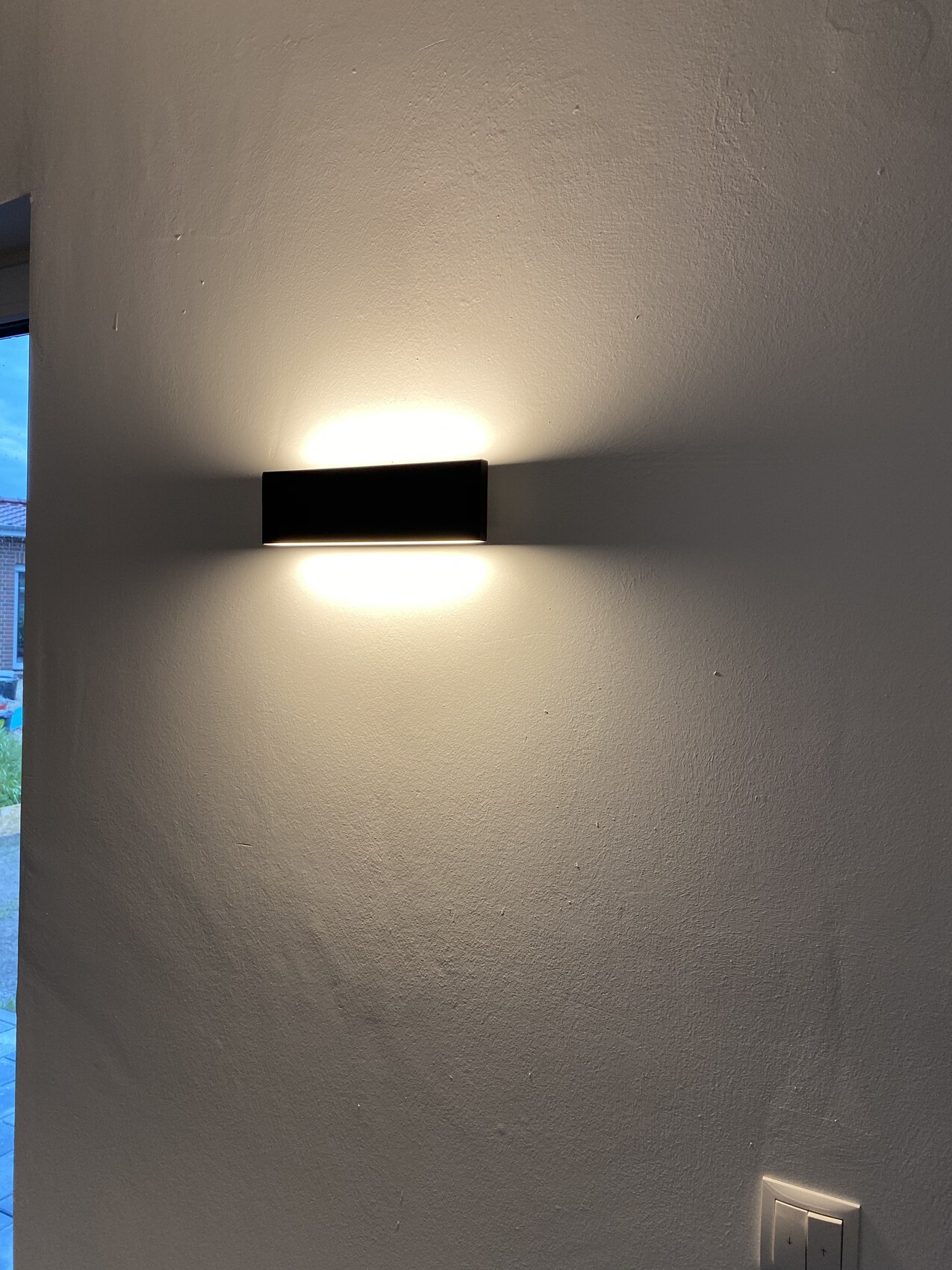 Schwarze flache rechteckige Wandlampe an weißer Wand, leuchtet oben und unten.