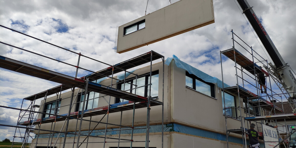 Baugerüst um ein modernes Haus; Kran hebt ein vorfabriziertes Hausteil-Modul aufs Obergeschoss.