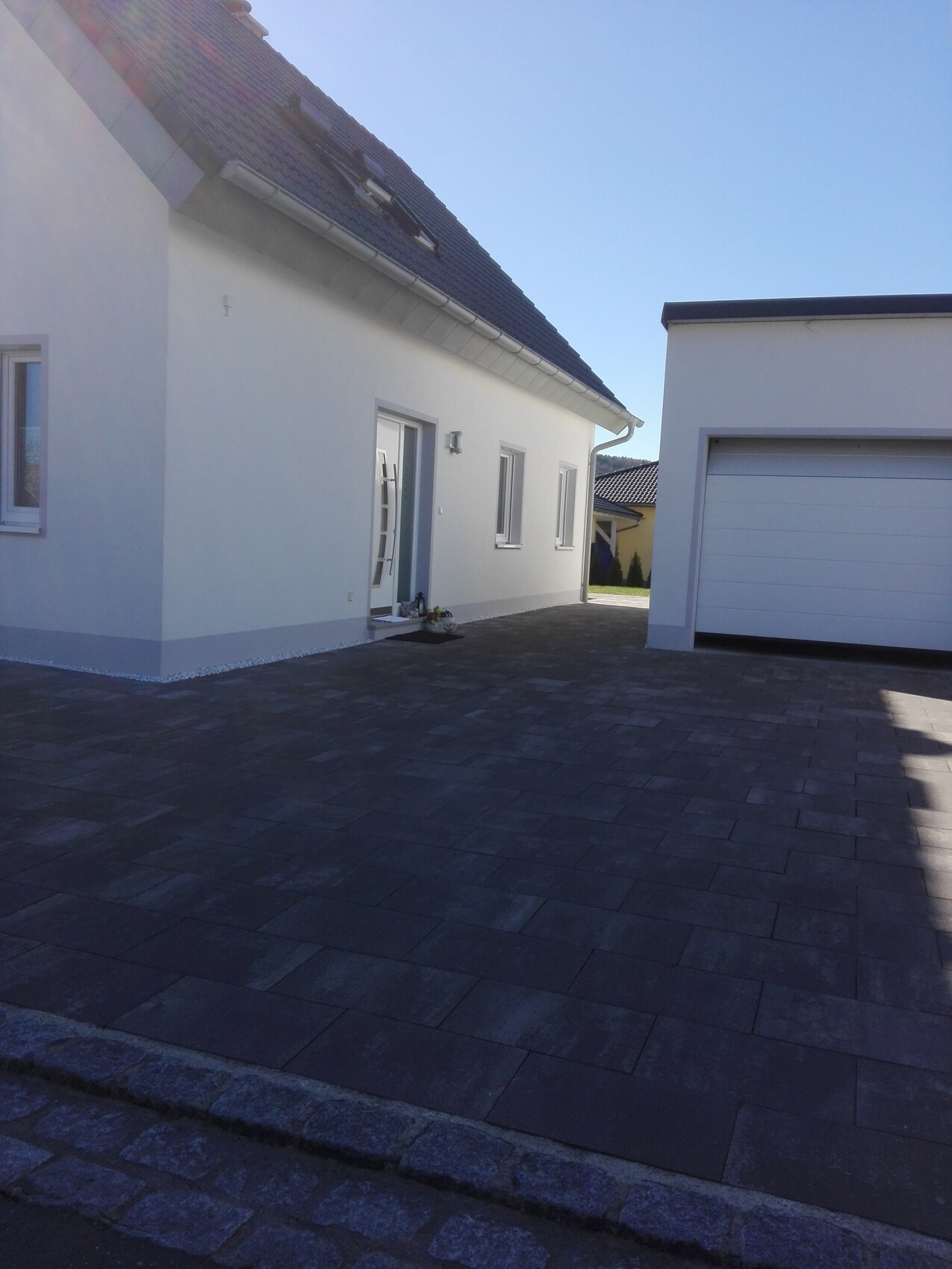 Modernes weißes Einfamilienhaus mit dunkler Pflaster-Einfahrt; rechts steht eine Garage.