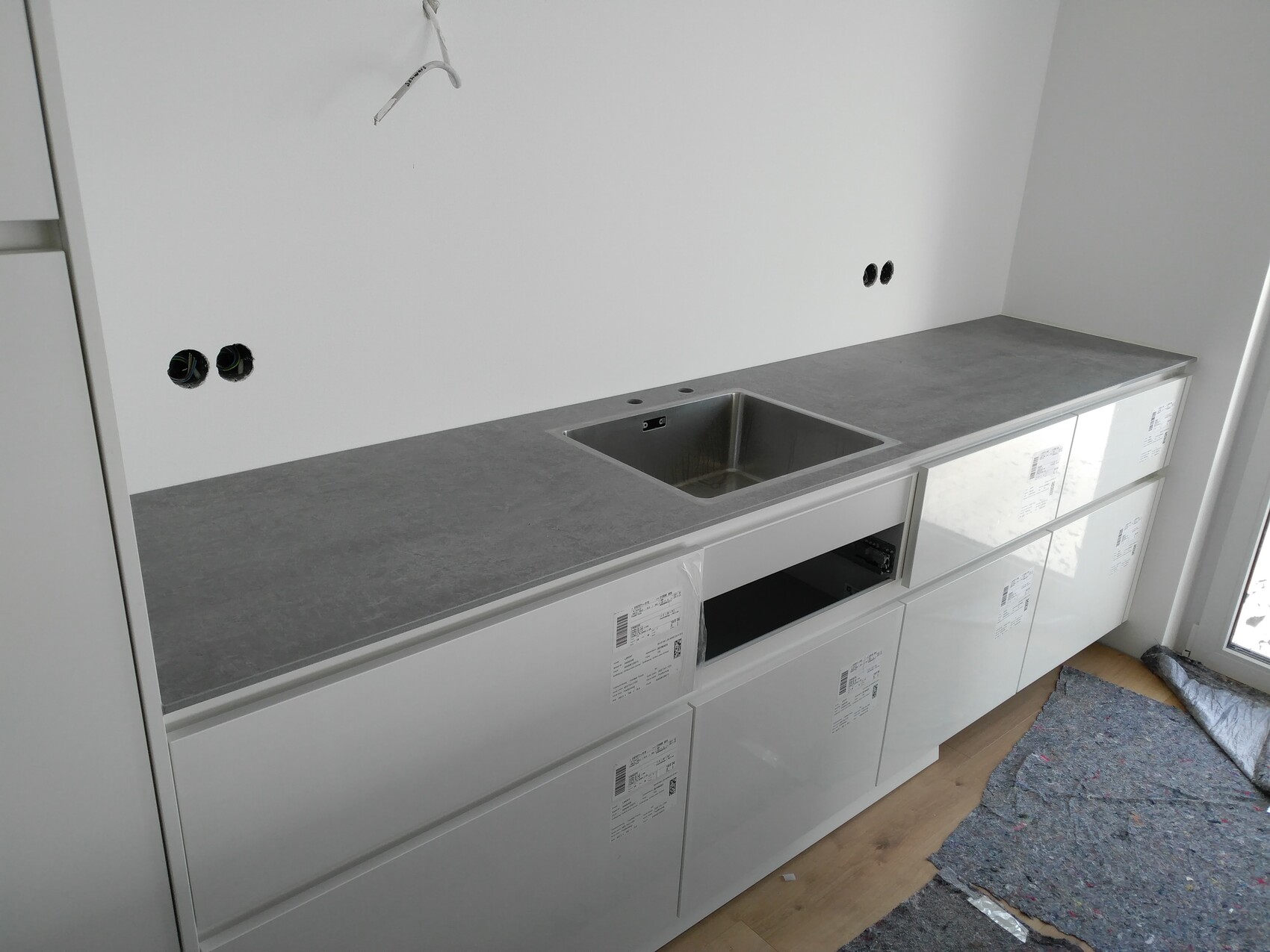 Küche in Bauphase: weiße Schränke, graue Arbeitsplatte, Spüle, Wand mit Steckdosenlöchern.