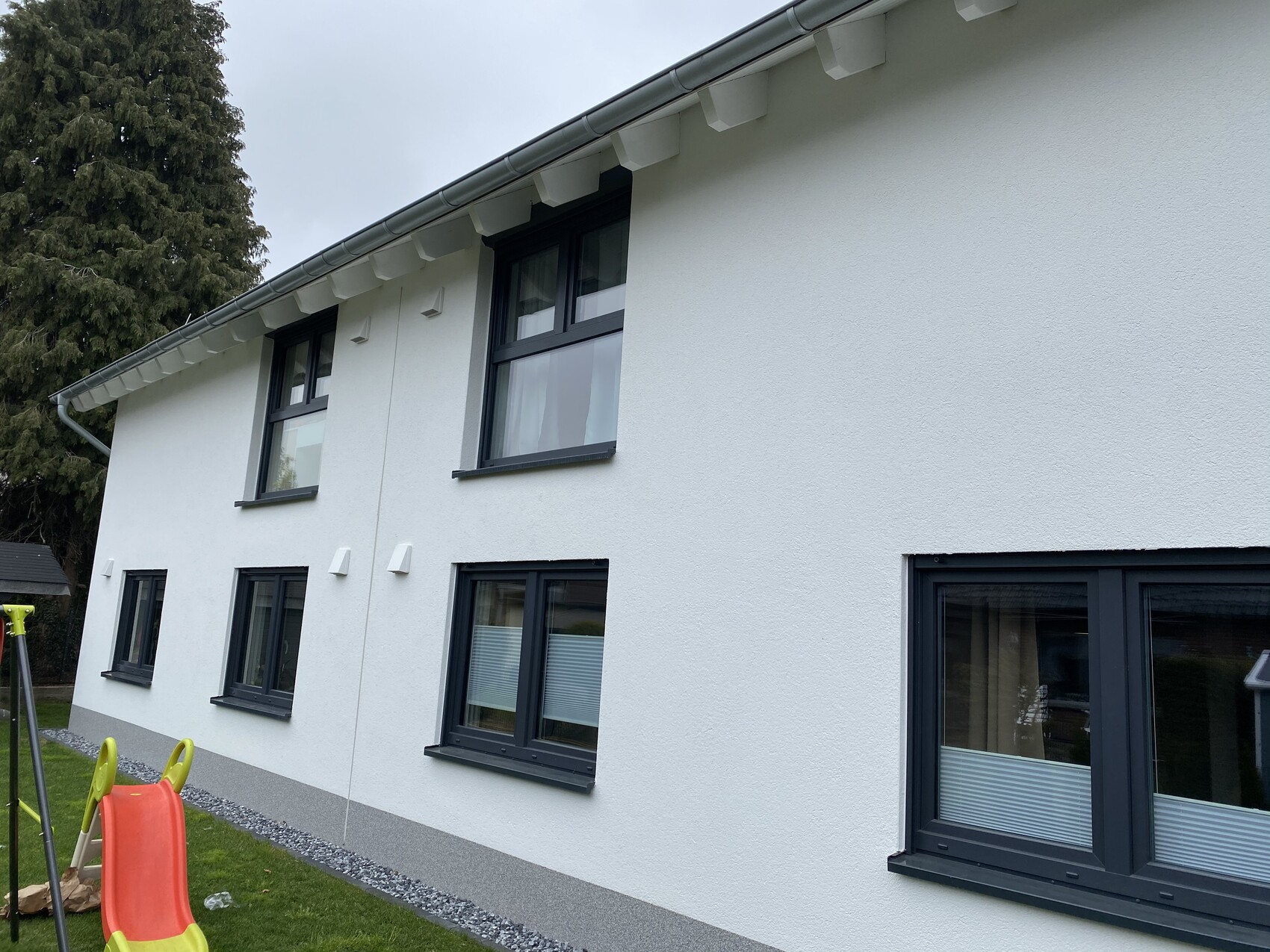 Moderne weisse Hausfassade mit dunklen Fenstern, Garten und Spielgeraet im Vordergrund