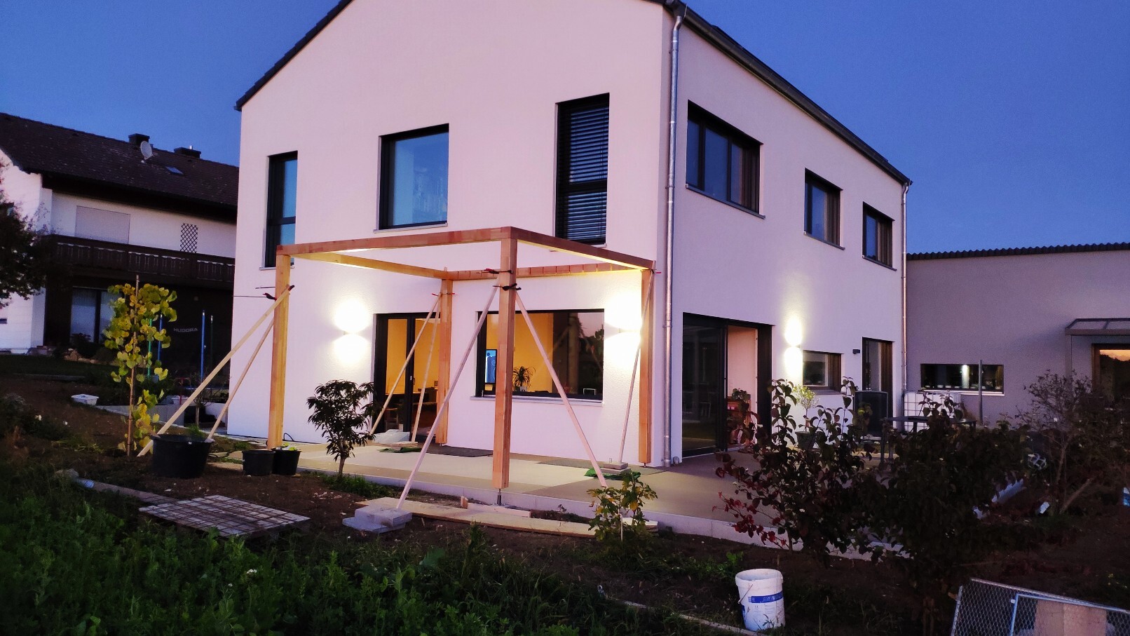 Modernes weißes zweistöckiges Haus mit Holz-Pergola im Hof; Abendlicht und Gartenbeleuchtung.