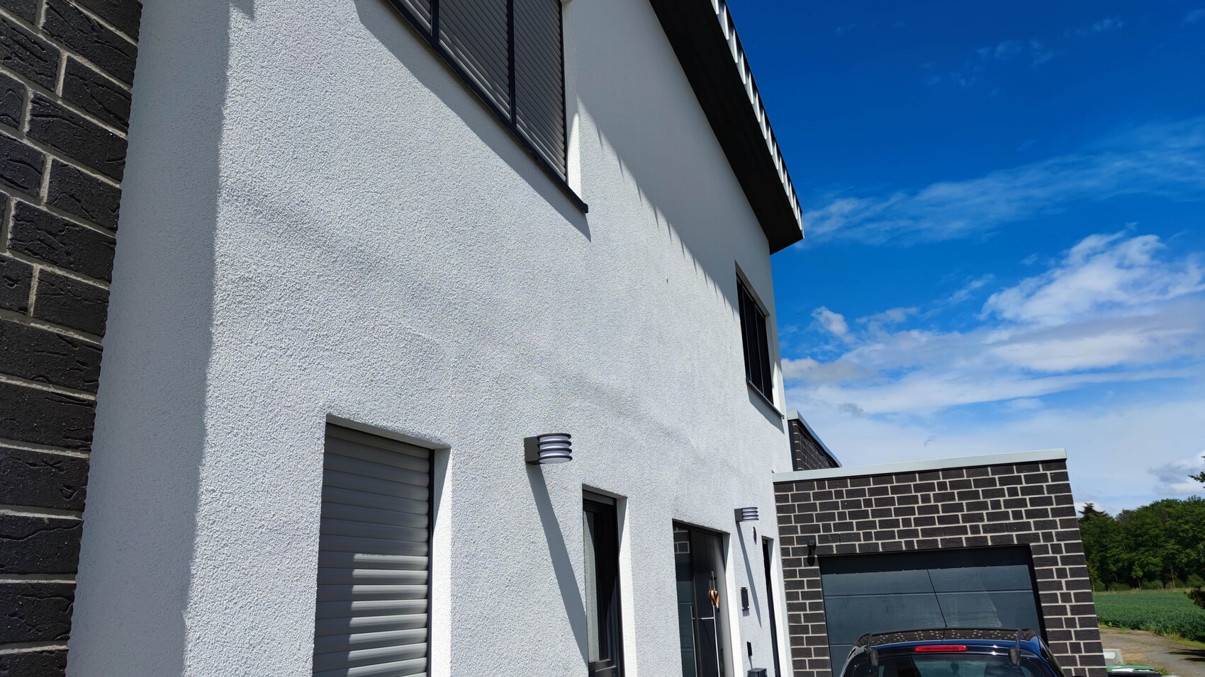 Weiße Hausfassade mit Rollläden, Außenlampen und Garage vor blauem Himmel.