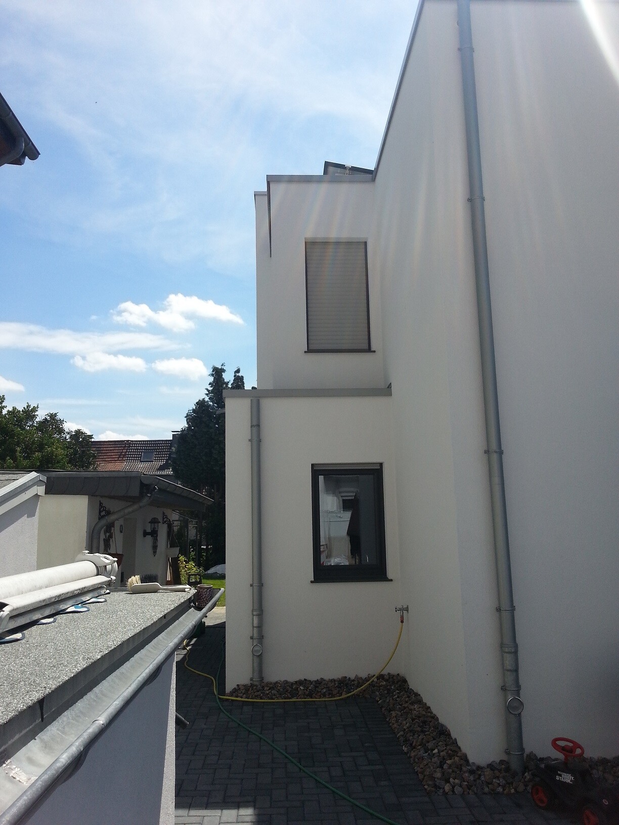Weiße moderne Gebäudefassade mit Fenstern, Regenrinne und gepflastertem Weg im Sonnenschein.