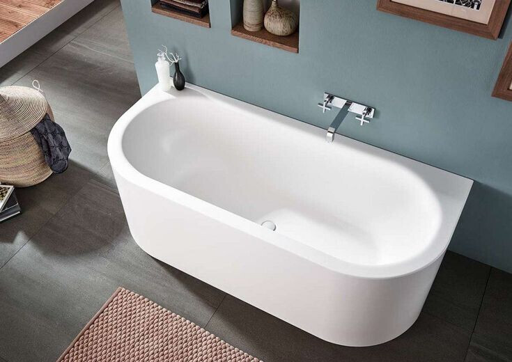 Weiße freistehende Badewanne vor blaugrüner Wand mit Ablagen und Deko.