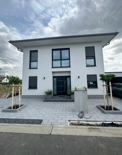 Moderne weiße Hausfront mit dunkler Eingangstür, gepflastertem Vorhof und Pflanzkübeln.
