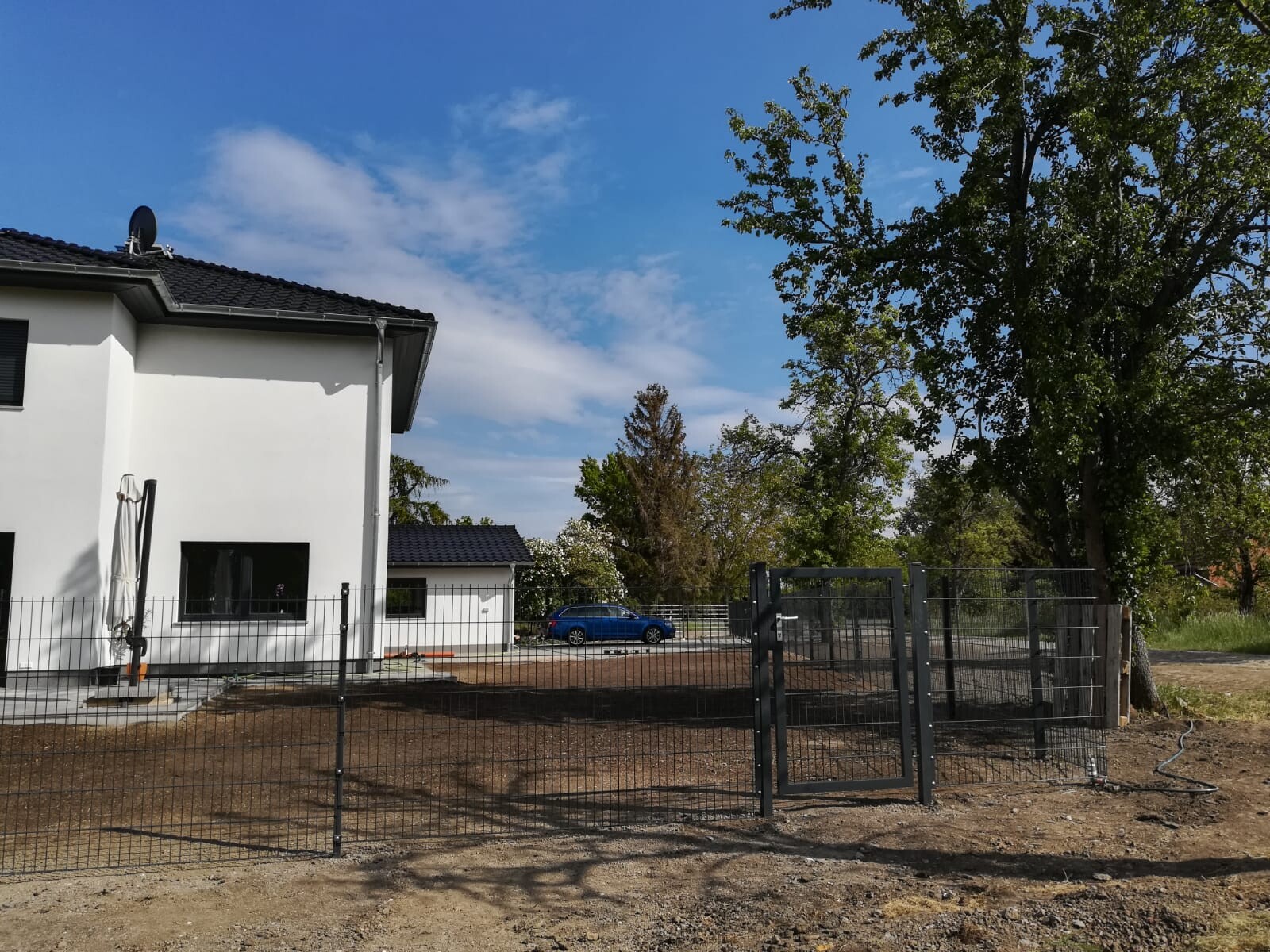 Weißes Haus mit schwarzem Dach, Gartenzaun und Tor; blauer Wagen im Hintergrund.
