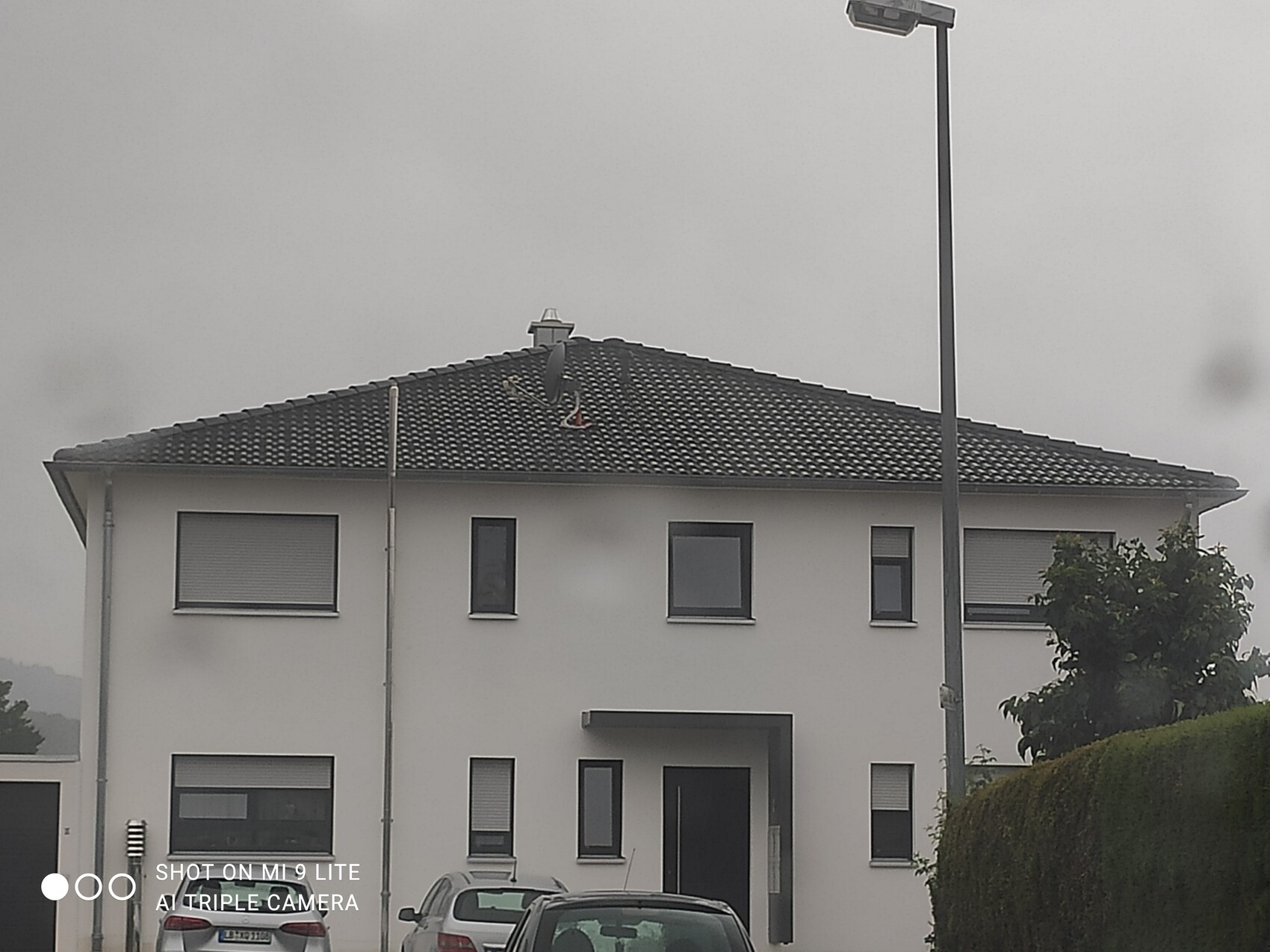 Weiße zweistöckige Wohnhausfront mit dunklem Ziegeldach, zwei Autos vor dem Eingang, Lampe rechts.