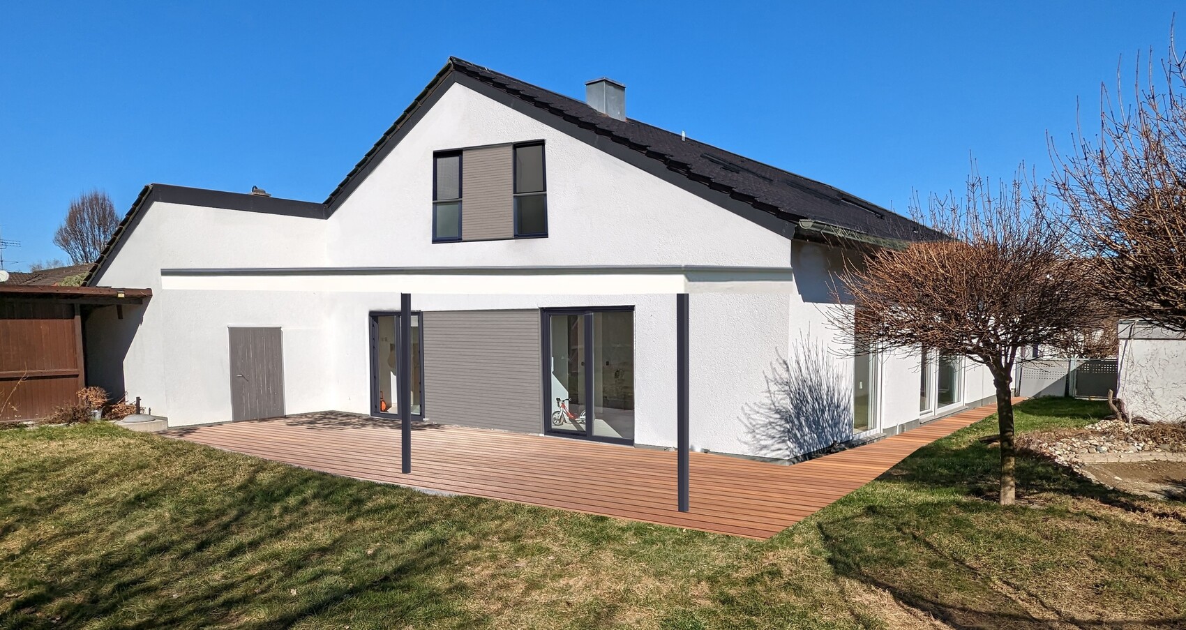 Weiße Hausfront mit dunklem Satteldach, Glasfronten und Holzterrasse vor Garten.