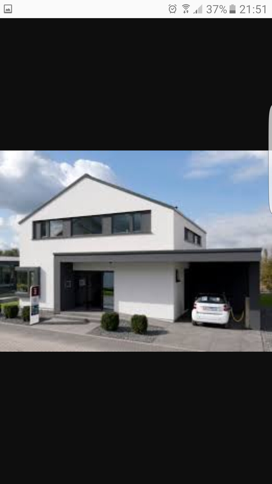 Modernes weißes Haus mit Carport, geparktem Auto und Garten vor blauem Himmel.