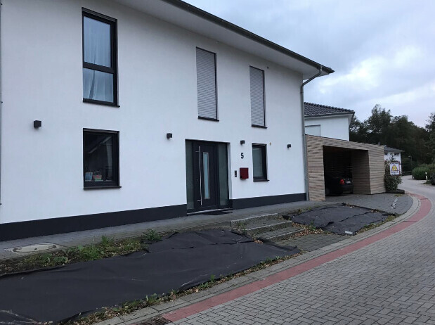 Weiße zweistöckige Hausfront mit dunkler Eingangstür, Rollläden und Carport rechts.