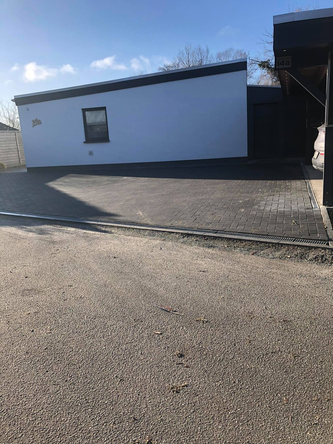 Weiße Hausfassade mit Fenster, gepflastertem Hof und Carport rechts mit Auto.