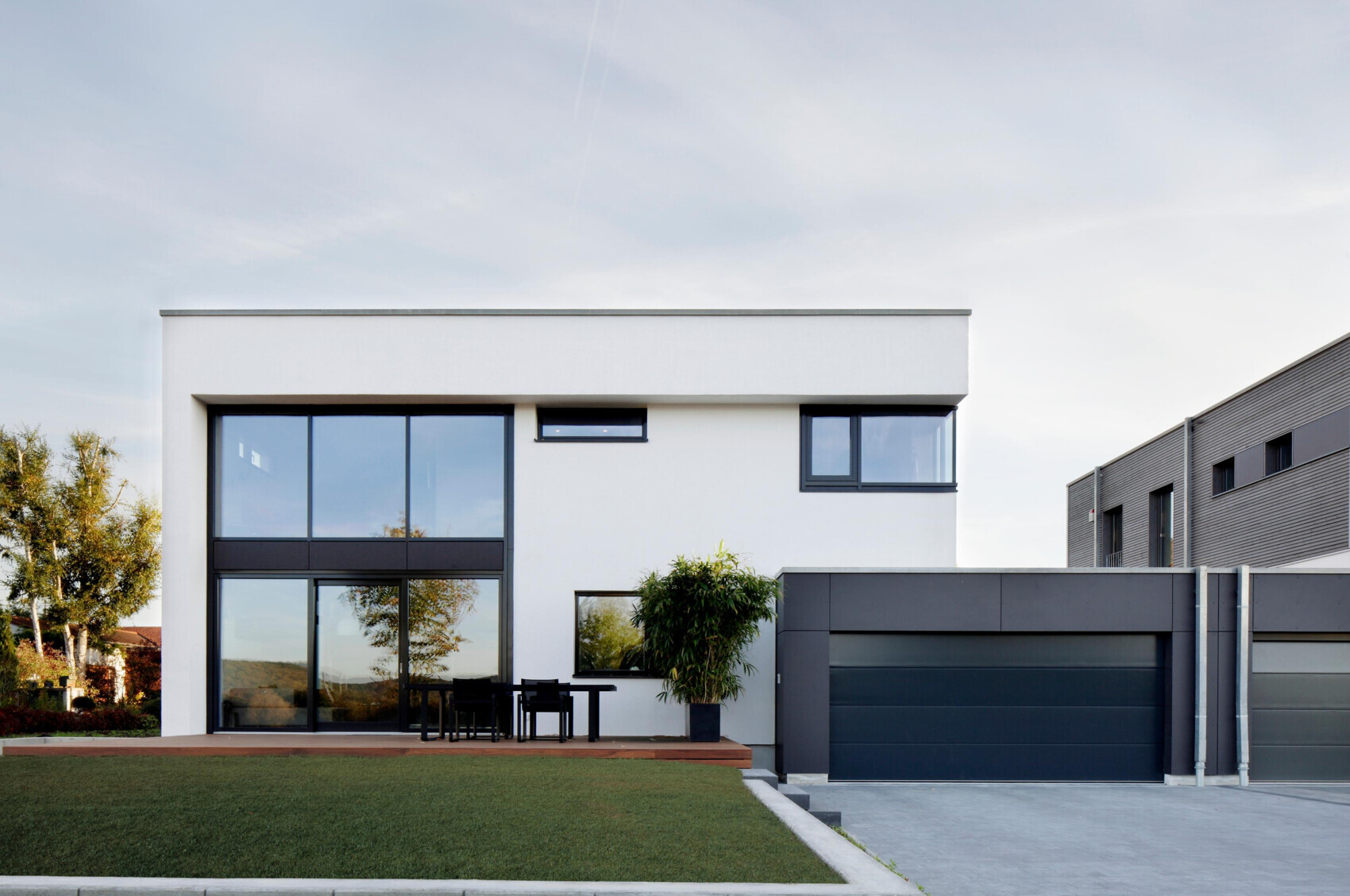 Moderne weiße Villa mit großen Glasfronten, Terrasse mit Tisch und Stühle, dunkle Garage rechts.