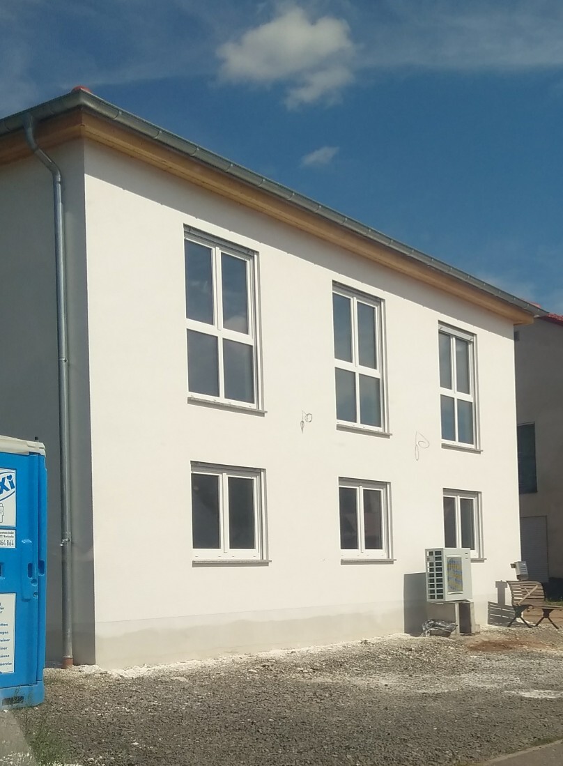 Weißes zweistöckiges Gebäude mit Fenstern; links Porta-Potty, rechts Außen-Klimagerät.