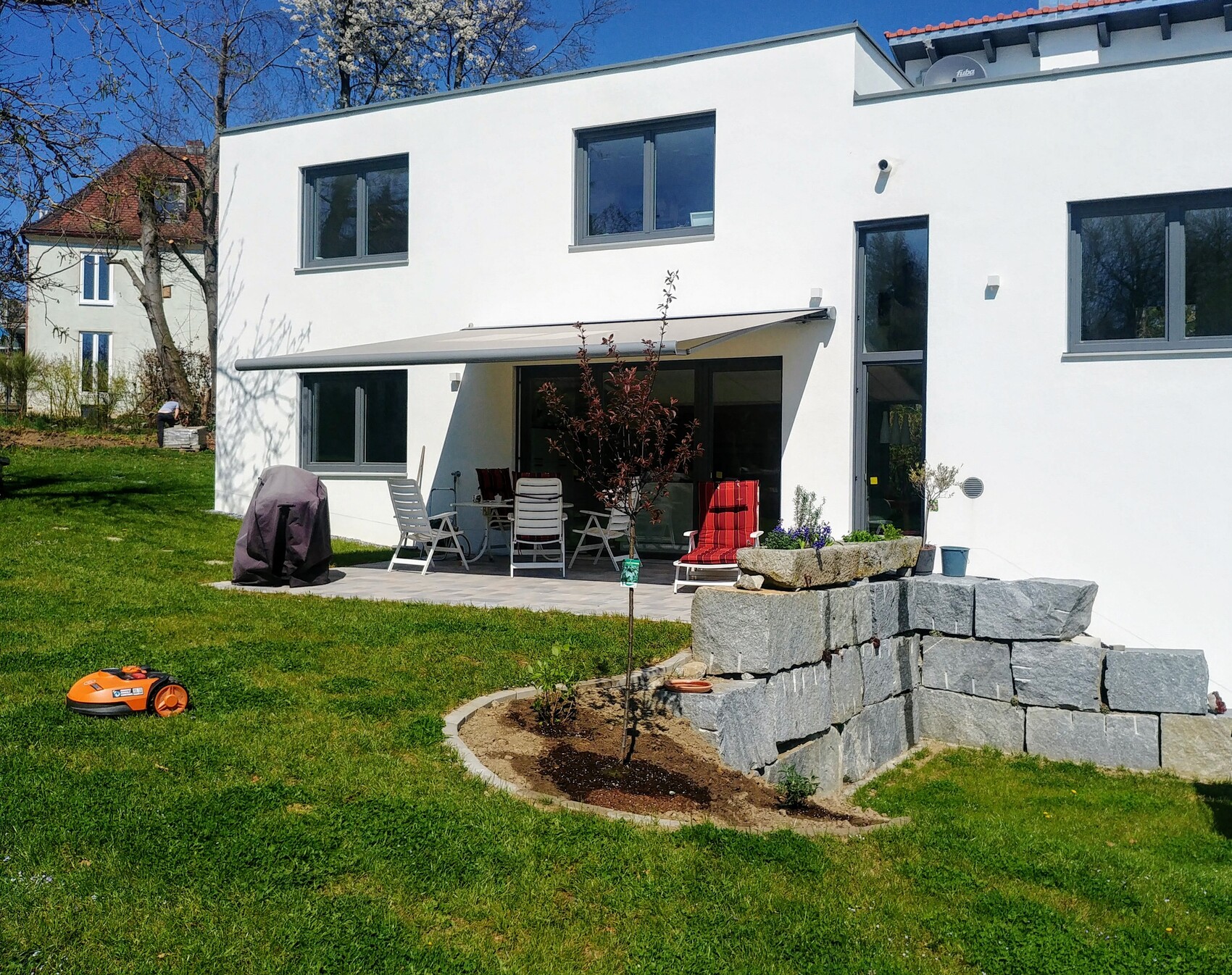 Weiße moderne Villa mit Terrasse, Sitzgruppe, abgedecktem Grill, Steinmauer und Rasen.