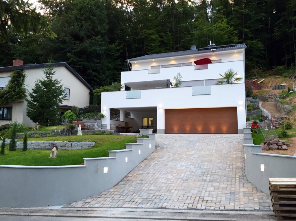 Moderne weiße Villa mit großer Holztor-Garage, gepflasterter Zufahrt und Garten am Hang.