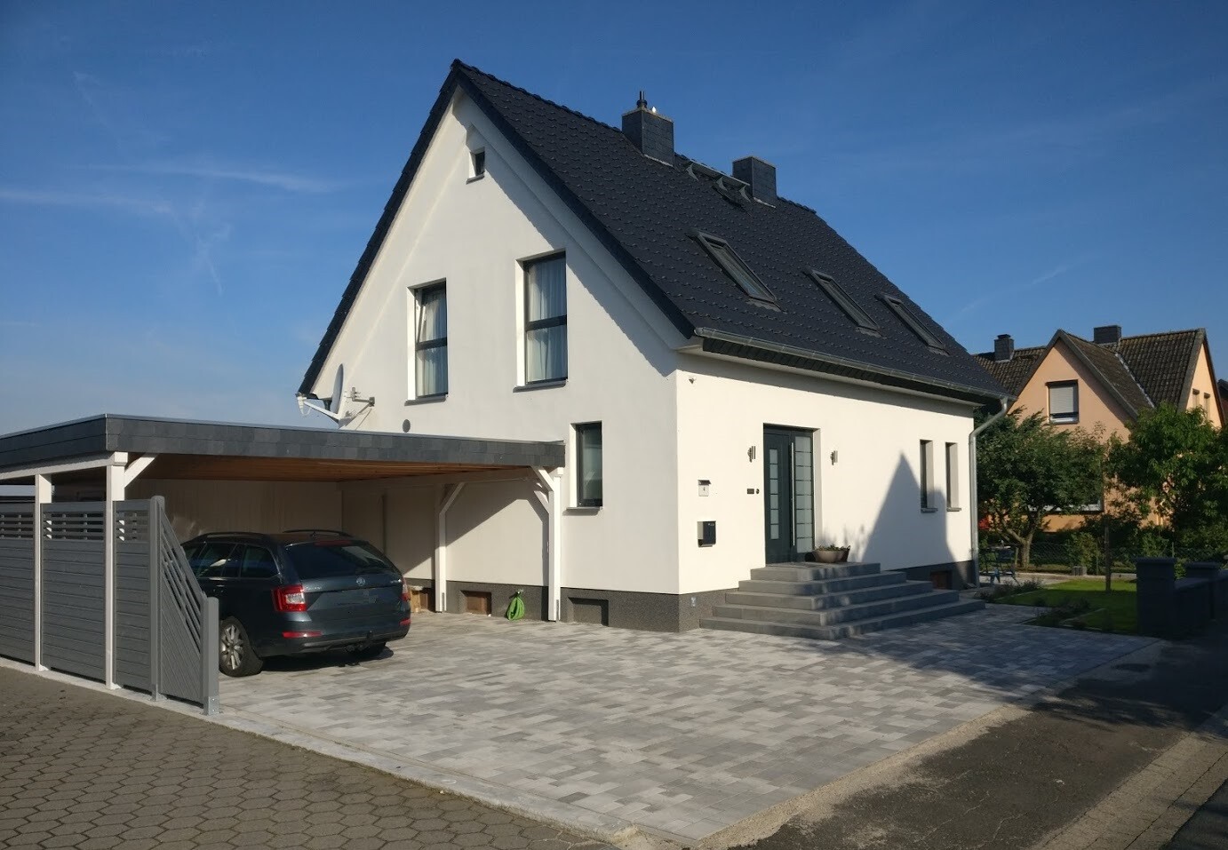 Helles Einfamilienhaus mit dunklem Satteldach, Carport mit Auto und gepflasterter Zufahrt.