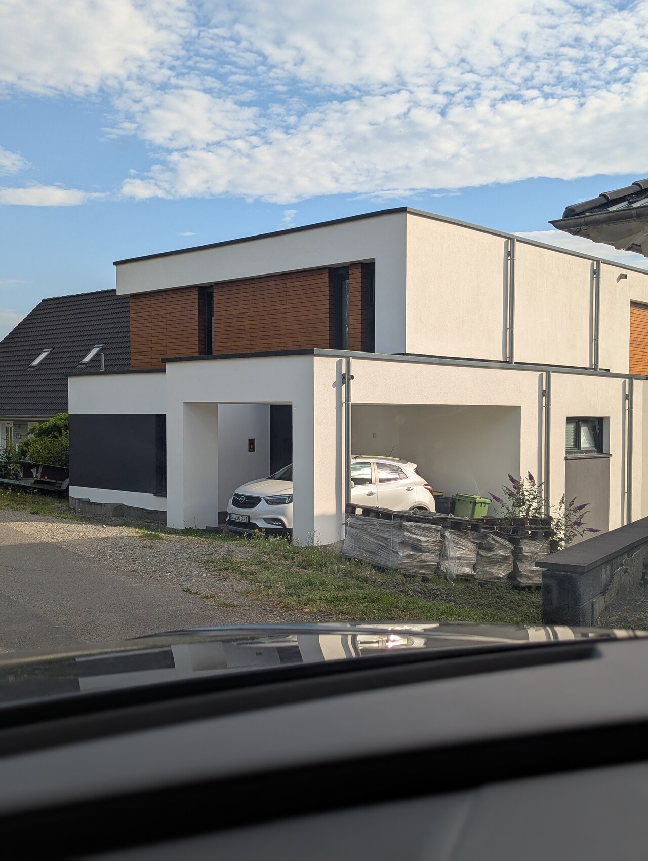 Moderne weiße Hausfront mit Garage; weißes Auto im Carport, daneben Paletten und Mülltonnen.