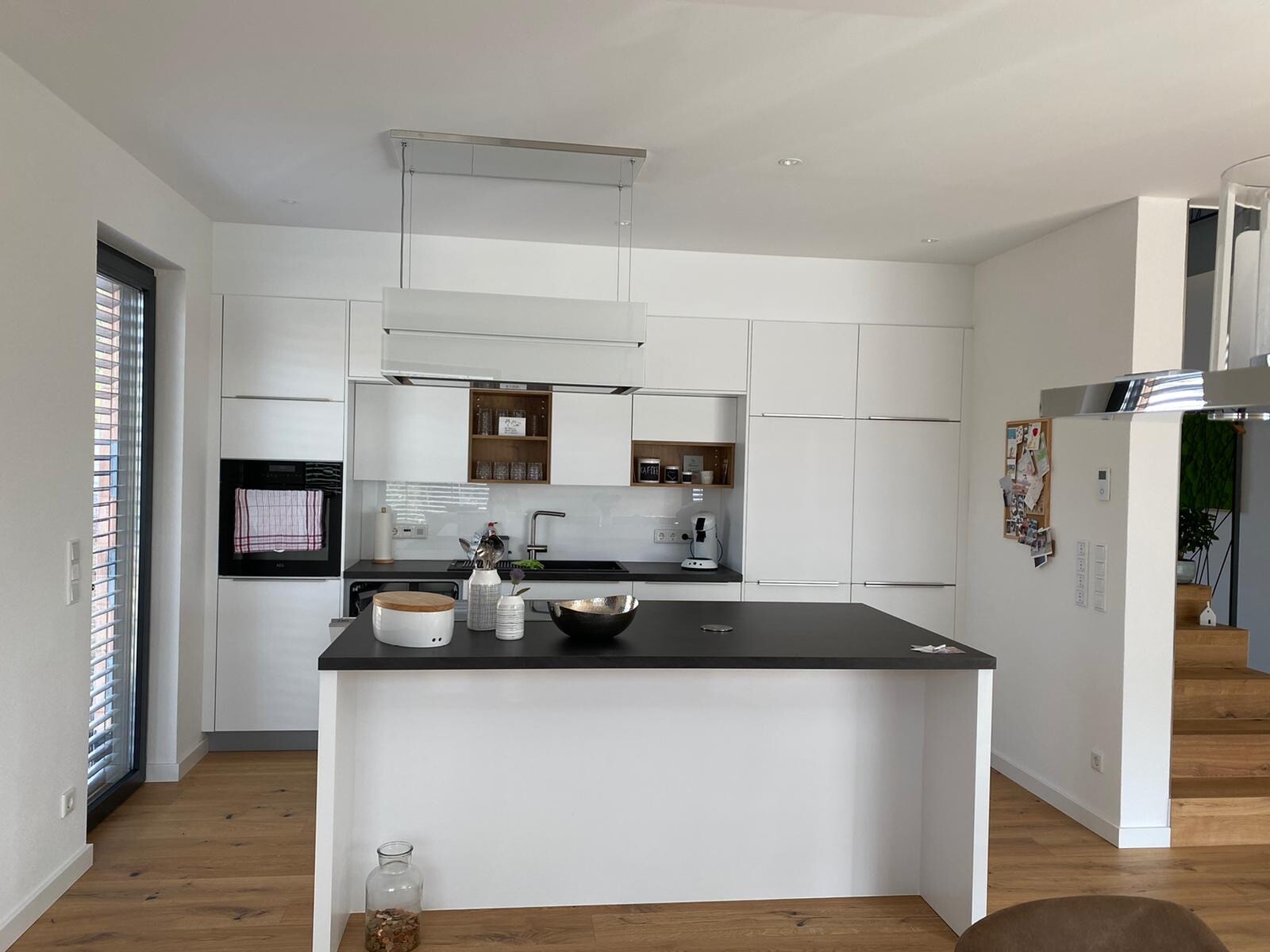 Moderne weiße Küche mit Insel, schwarzer Arbeitsplatte, Holzboden und Treppenbereich.