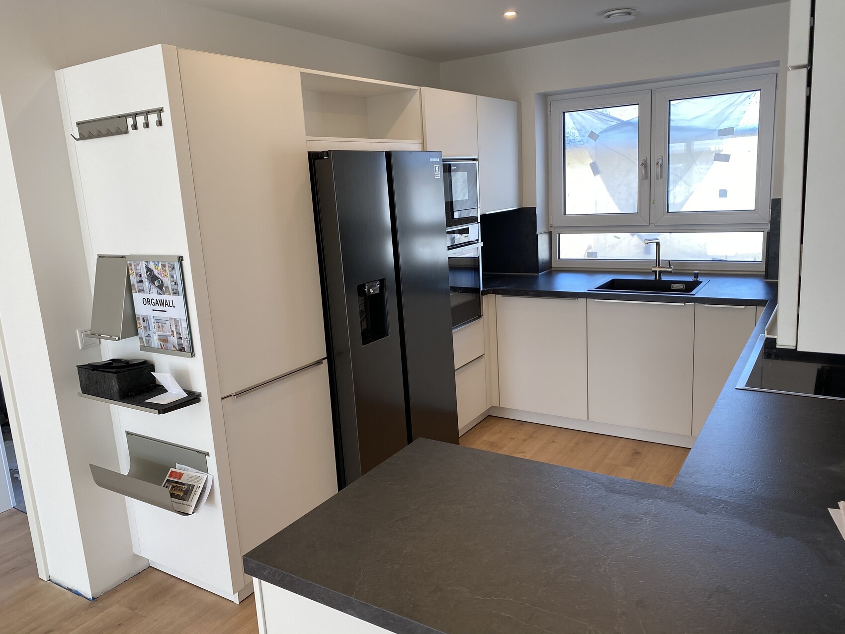 Moderne weiße Küche mit schwarzer Arbeitsplatte, Edelstahlkühlschrank, Ofen, Spüle vor Fenster.
